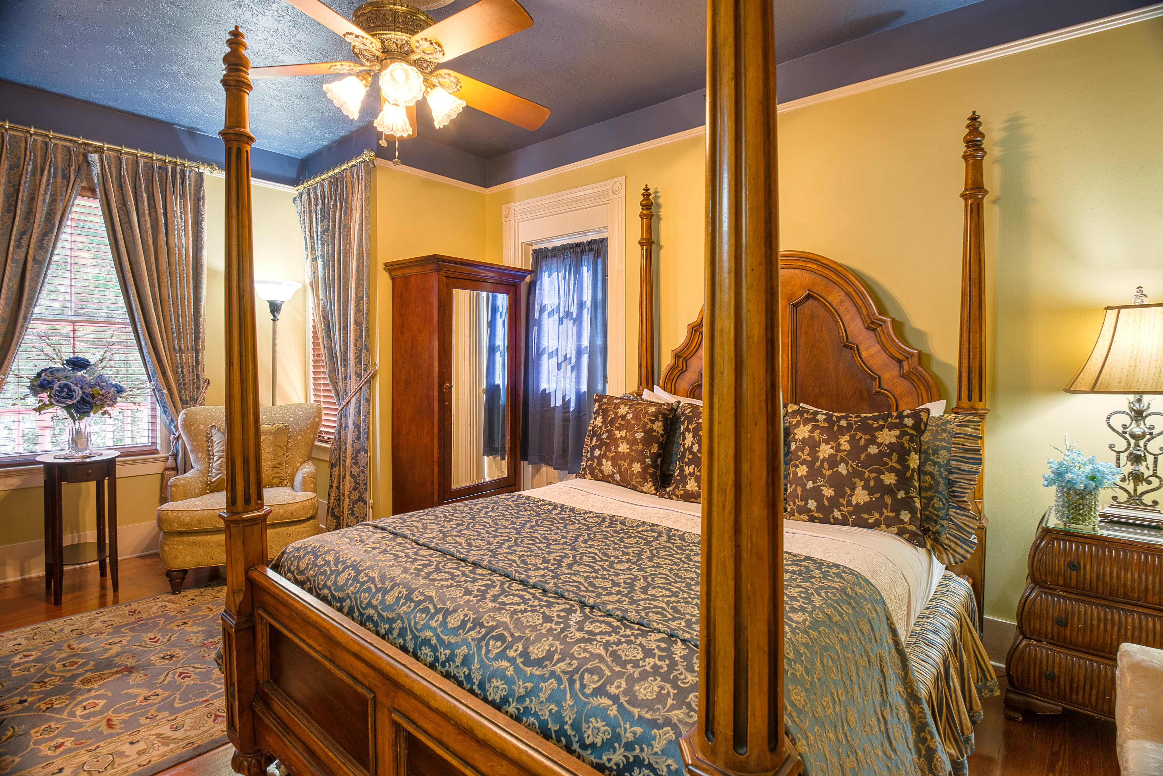 A spacious room with a four-poster bed, a dark wood dresser, a sitting area with a chair and a lamp, and a sunlit window.