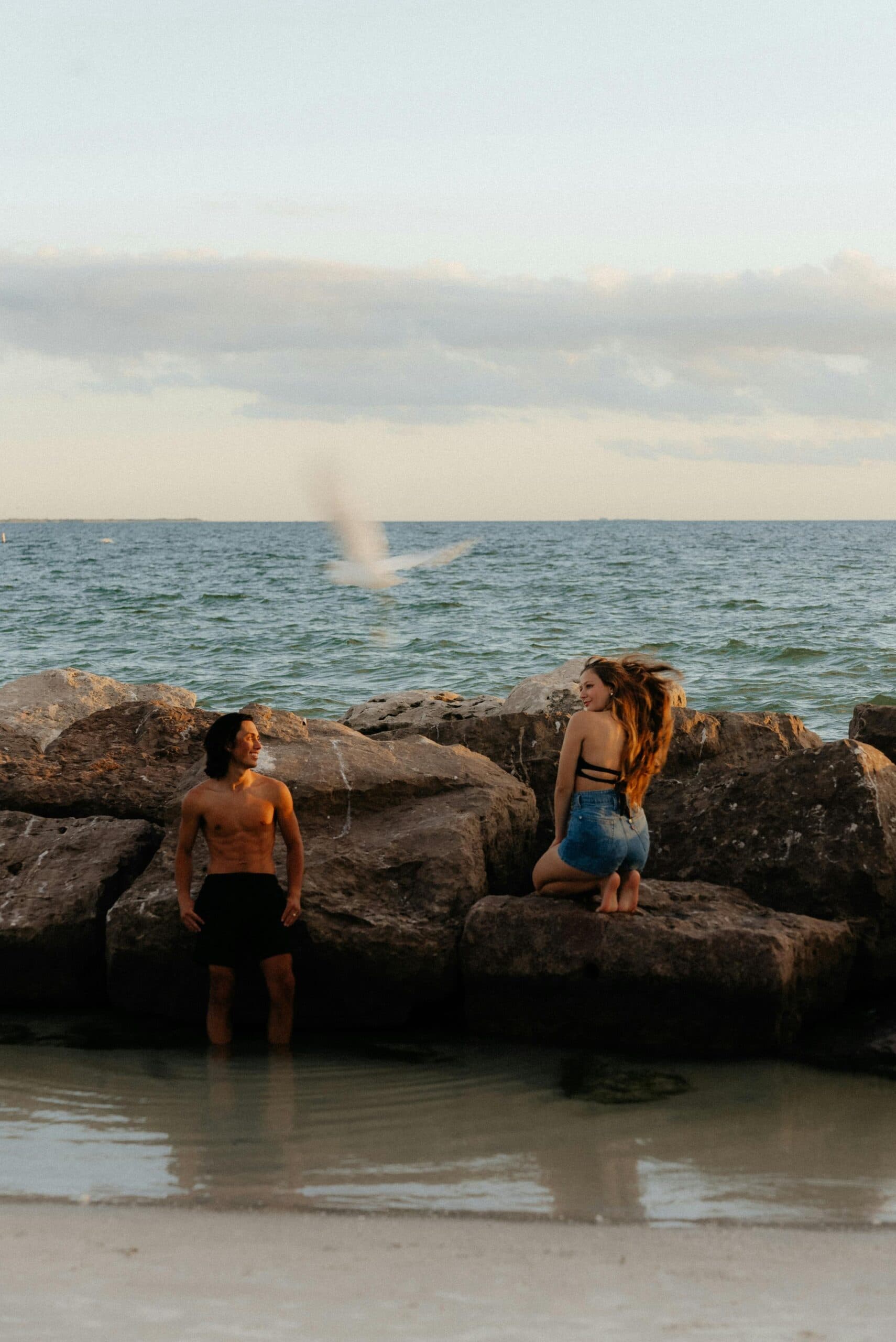 A man and woman interact playfully by the water's edge besides large rocks on a beach. A man and woman interact playfully by the water's edge besides large rocks on a beach.