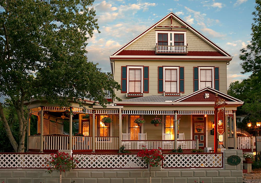 A charming two-story house with decorative trim, a porch, and lush greenery.