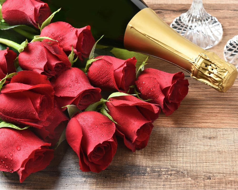 A bouquet of red roses next to a bottle of champagne on a wooden surface.