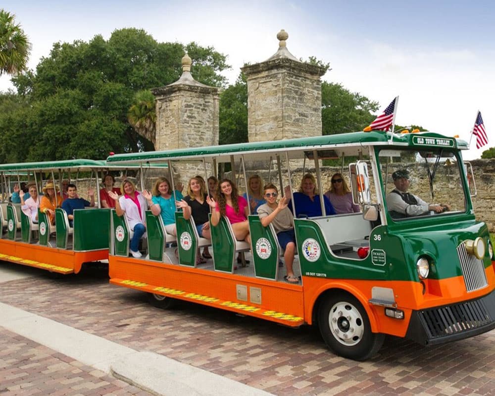 A tour trolley with smiling passengers waves at the camera in front of historic stone structures.