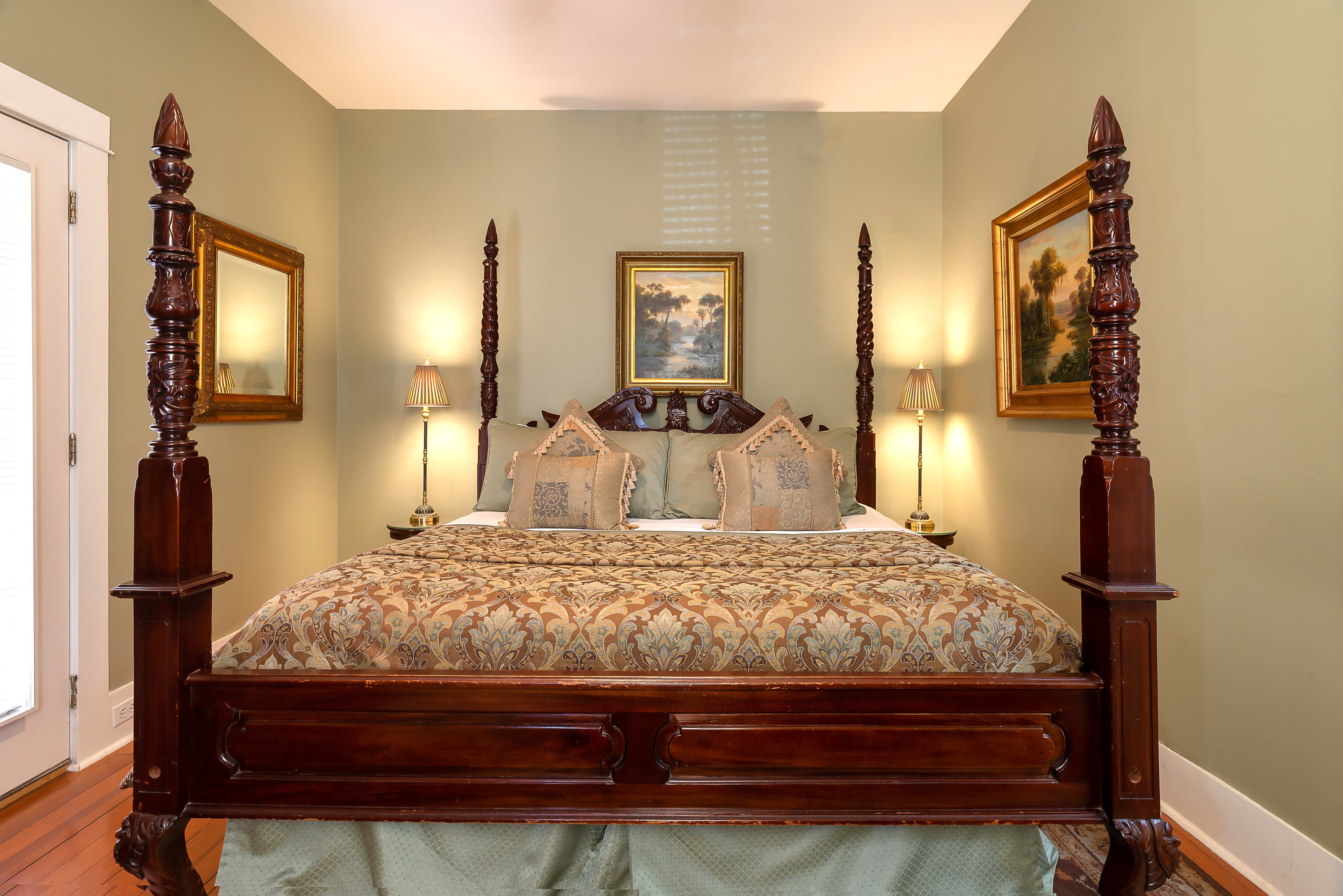 A traditionally styled bedroom with a dark wood four-poster bed, two framed pictures hanging on the wall, and two lamps on nightstands.