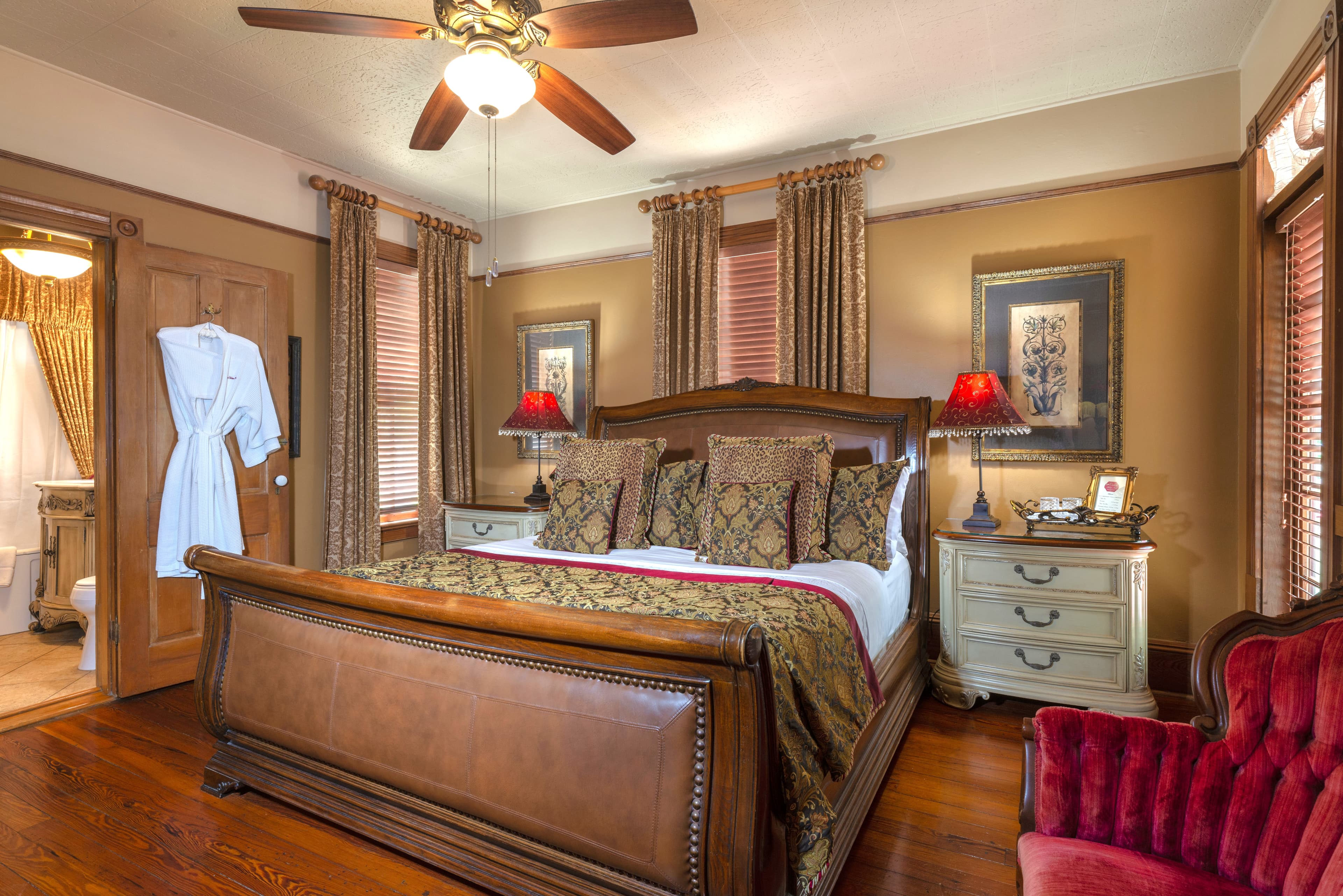 An elegant room with a sleigh bed, a dark wood dresser, a red armchair, and a bathroom with a shower and a sink. A framed picture hangs on the wall above the bed.