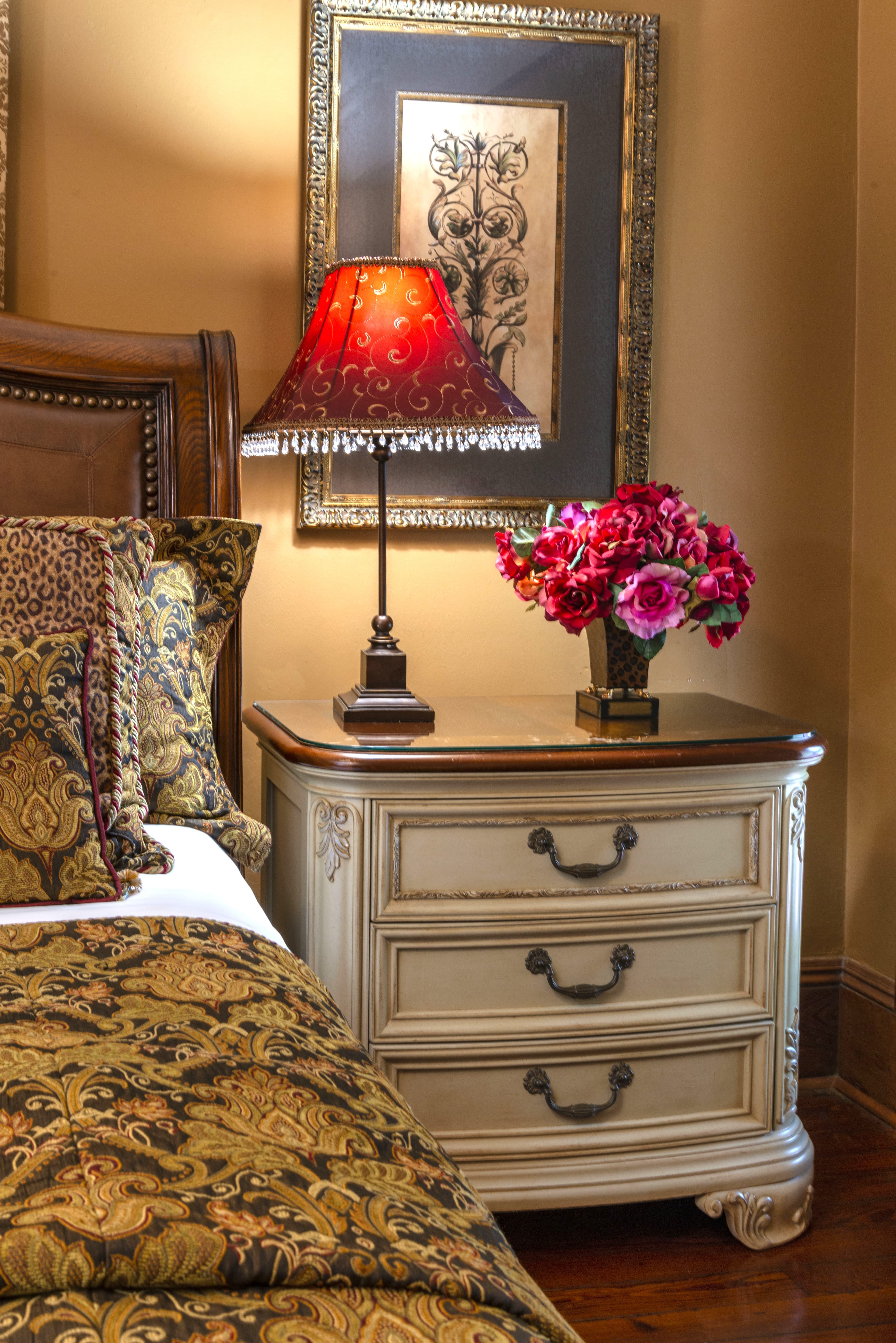 A romantic bedroom with a dresser nightstand topped with a table lamp and a vase with flowers next to a turned down bed.re hangs on the wall above the dresser.