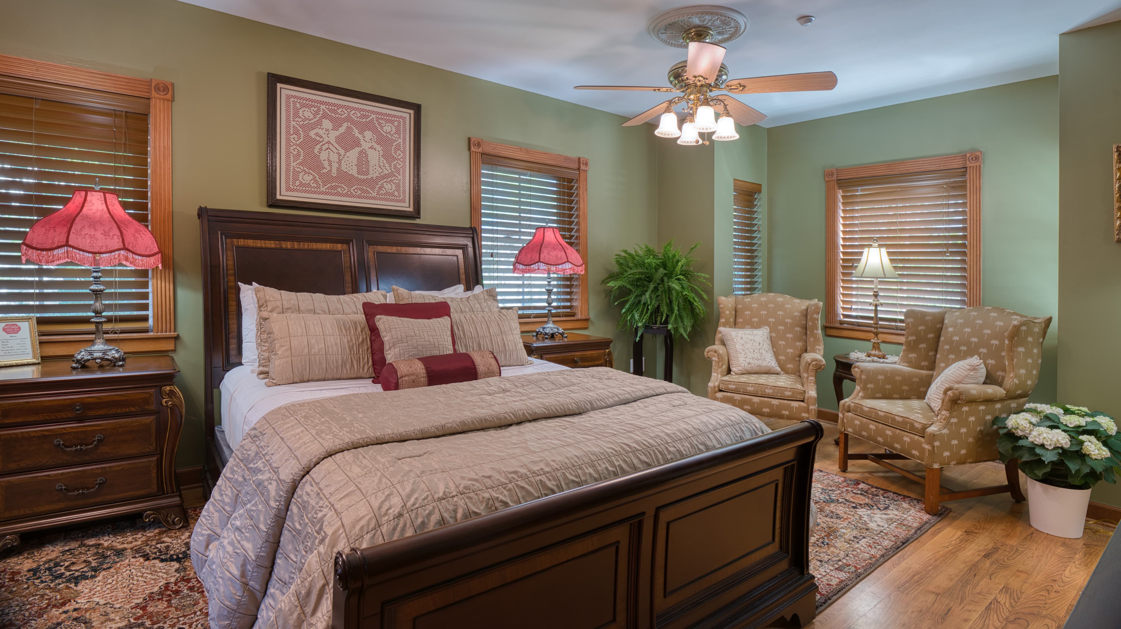 A traditionally styled bedroom with a dark wood bed frame, a sitting area with two armchairs, and two windows with wooden frames.