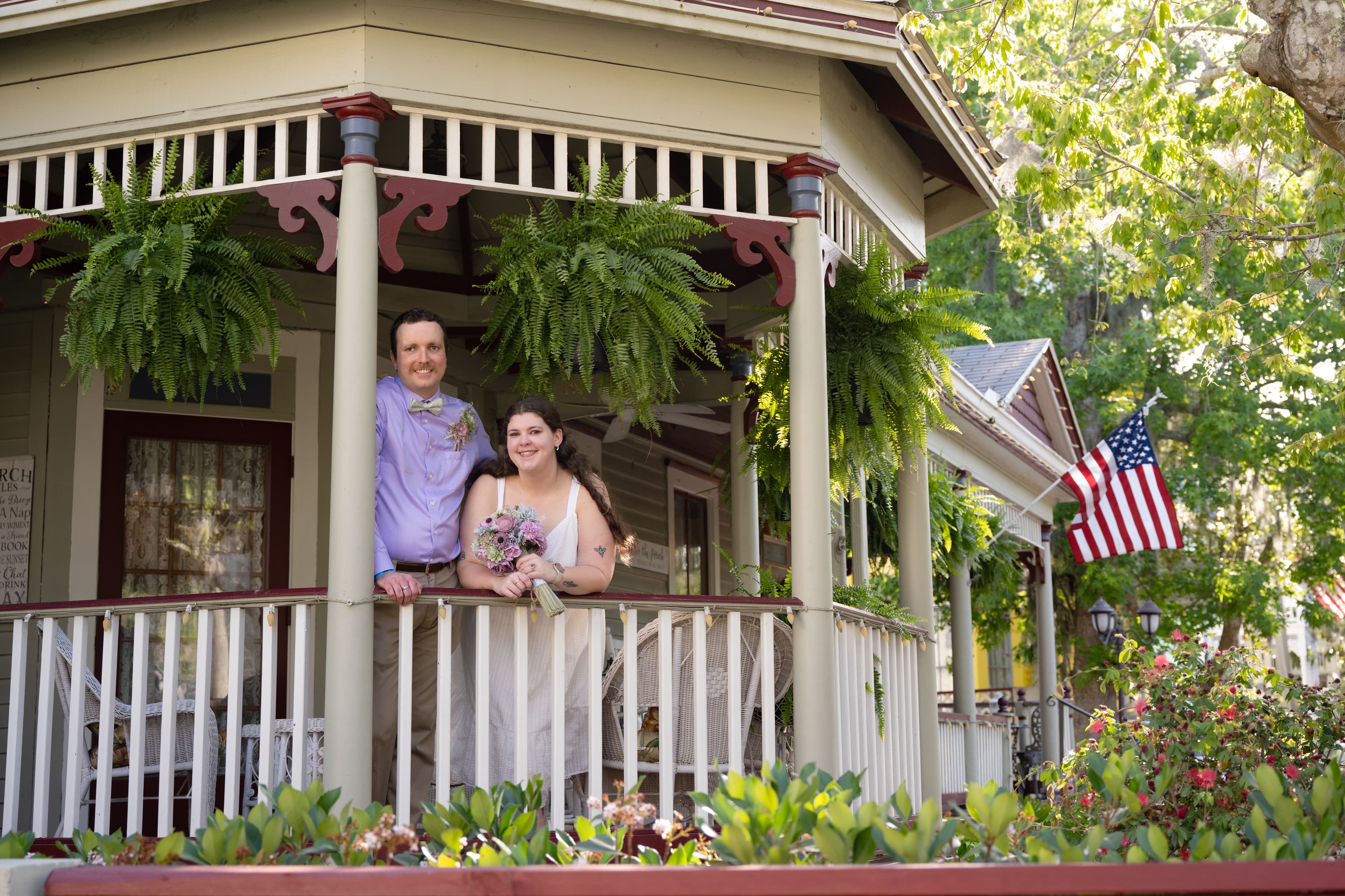 A couple smiles together on a porch adorned with ferns, holding a bouquet and surrounded by greenery.