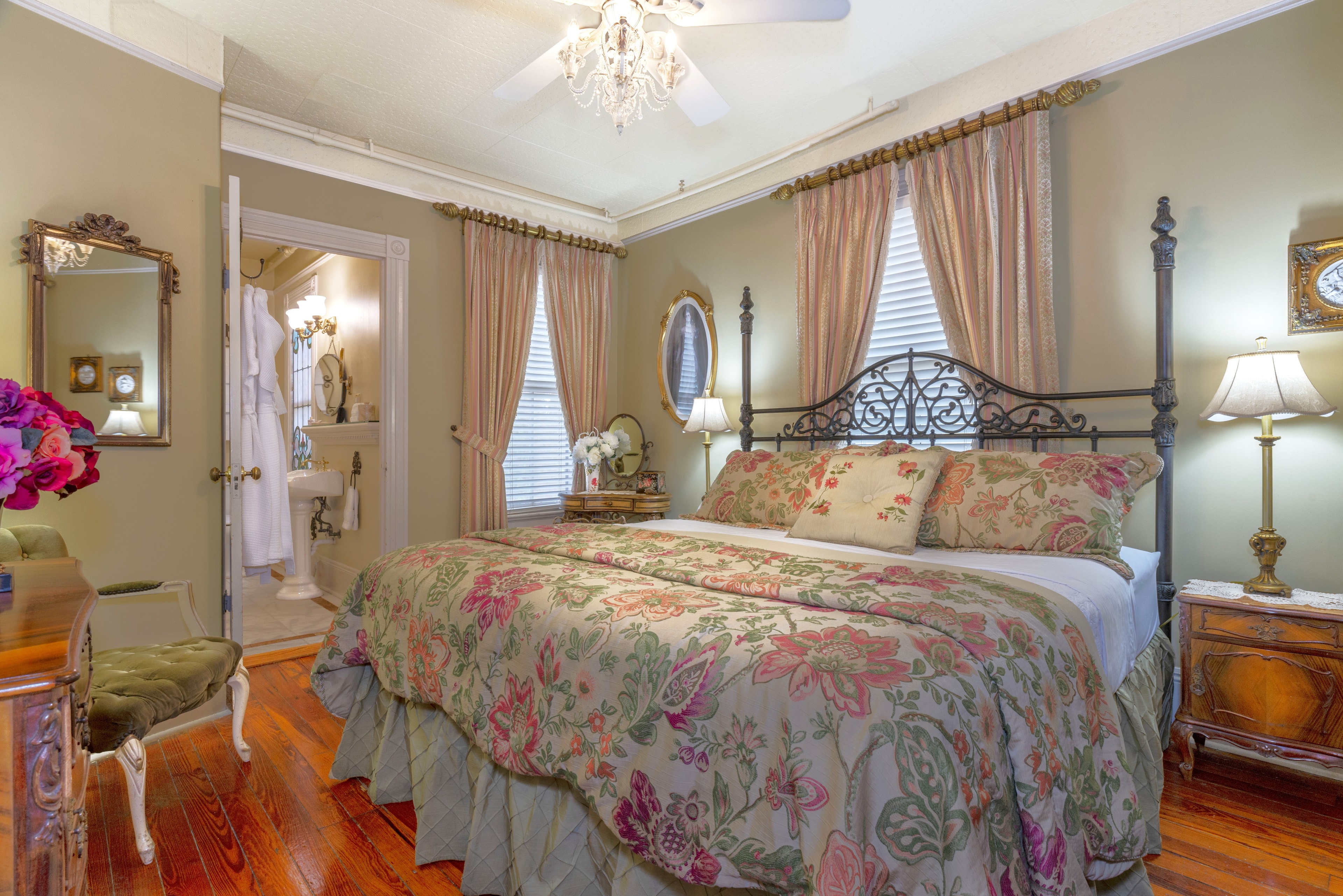 Interior view of a hotel room featuring a large bed with a patterned comforter and pillows, a dark wood dresser, a sitting area with a chair and a lamp, and a bathroom with a shower, a sink, and a mirror.