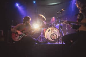 A three-member band performs on stage with colorful lights, featuring a guitarist, drummer, and bassist.