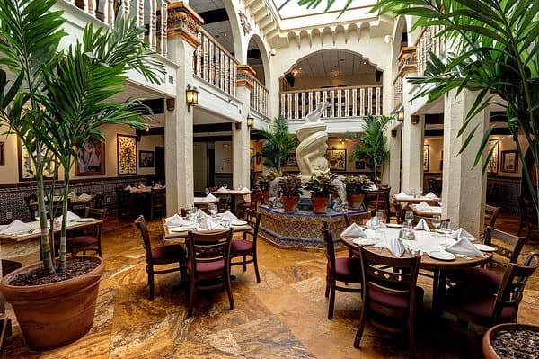 Elegant restaurant interior with potted palm trees and a central fountain surrounded by dining tables.