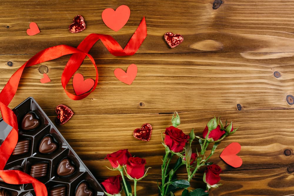 A box of chocolates, red roses, and decorative hearts on a wooden surface.