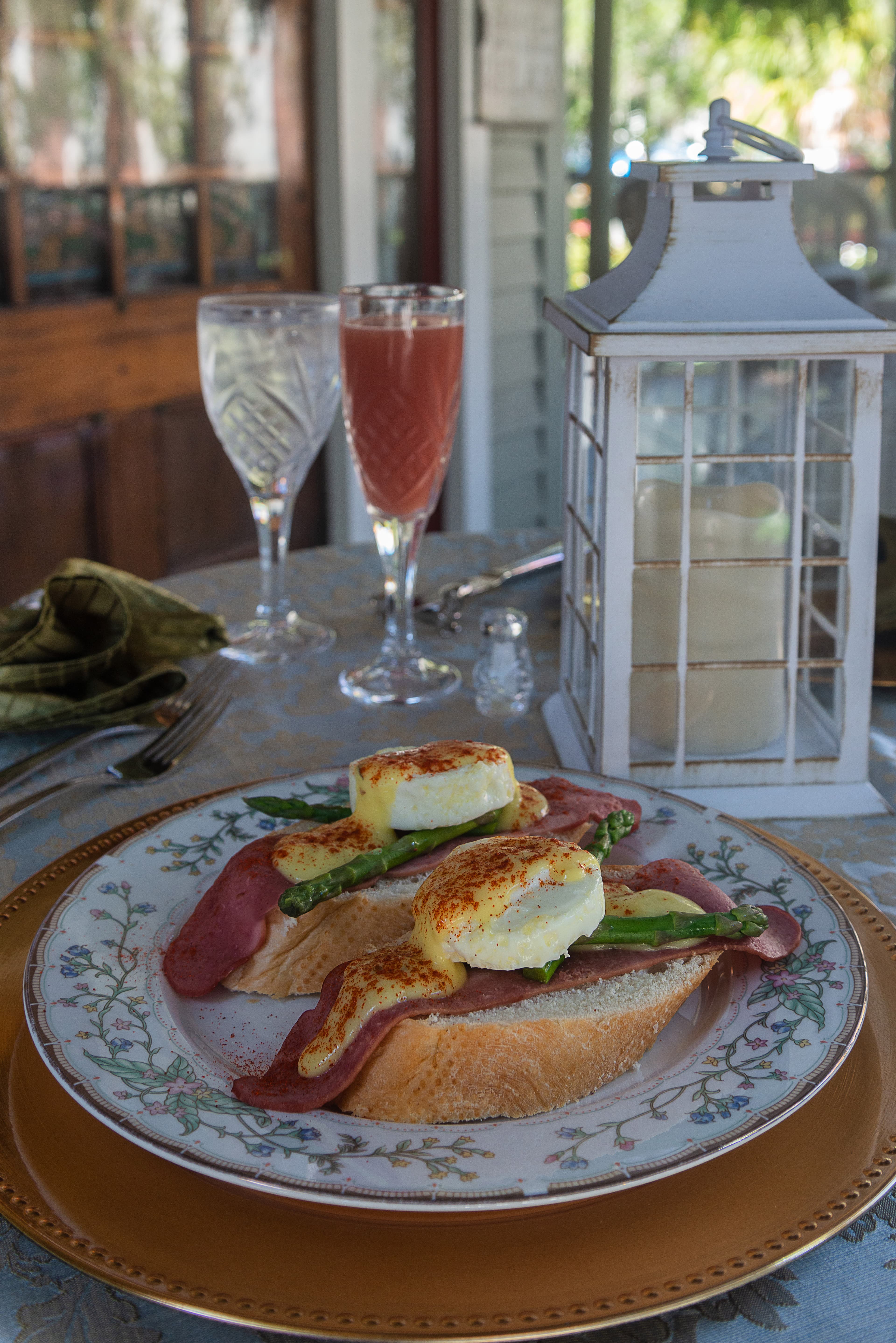 A beautifully arranged plate of eggs Benedict on toast with asparagus, accompanied by two elegant glasses of beverages and a lantern for ambiance.