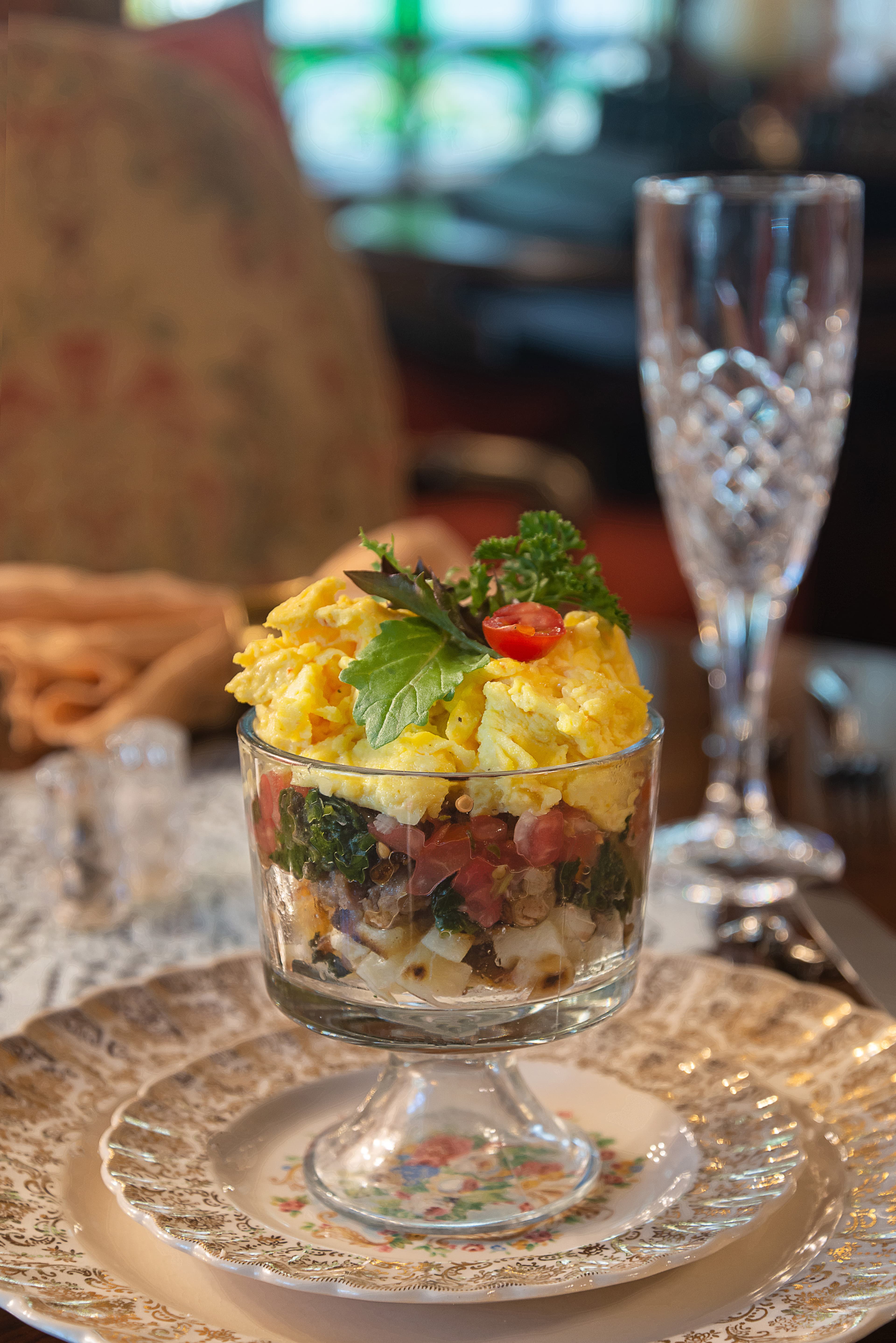 A layered salad of greens, tomatoes, and grains topped with fluffy scrambled eggs in a glass dish, served on an ornate plate.