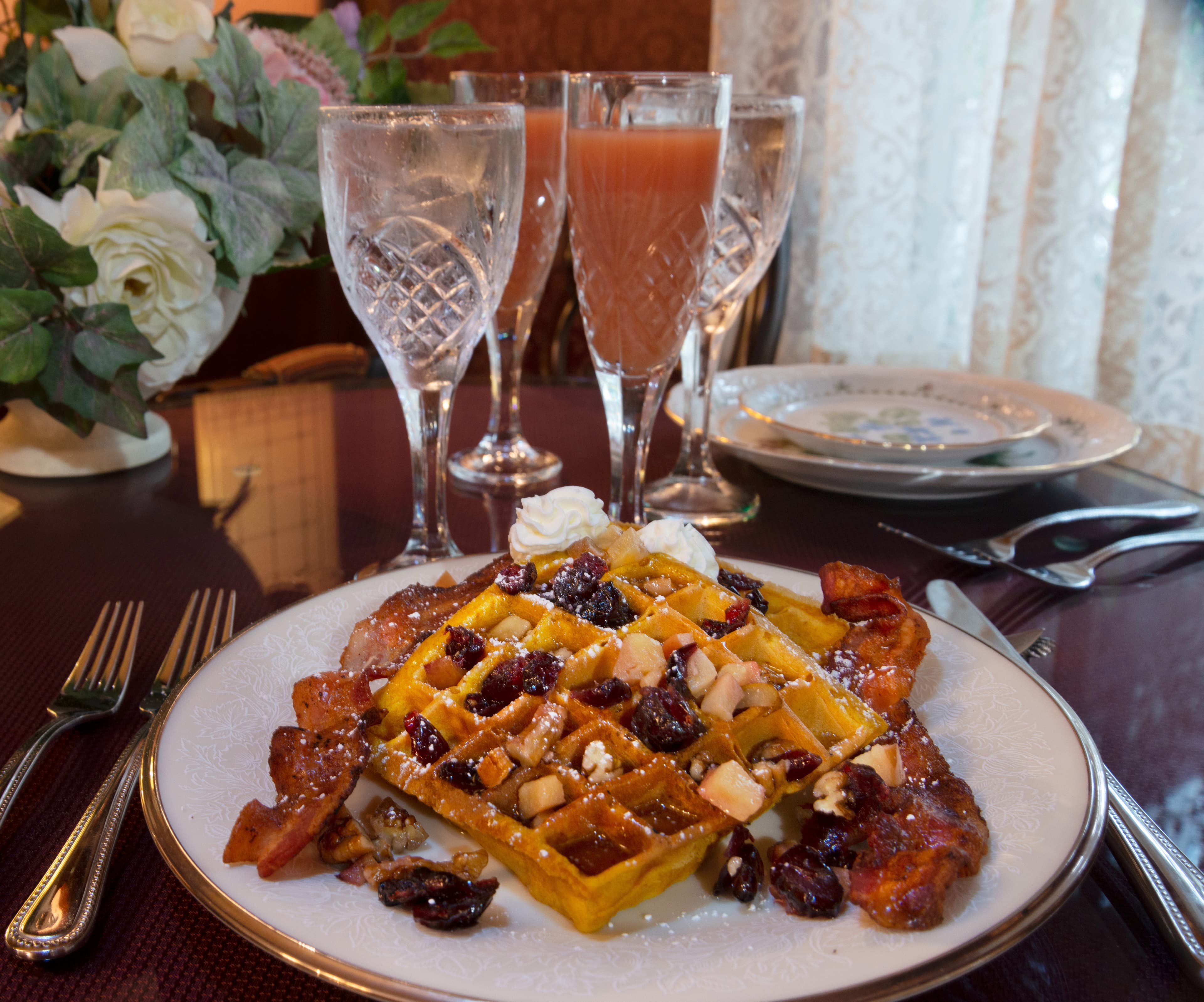 A plate of waffles topped with nuts, dried fruit, and whipped cream, accompanied by bacon and drinks in elegant glasses.
