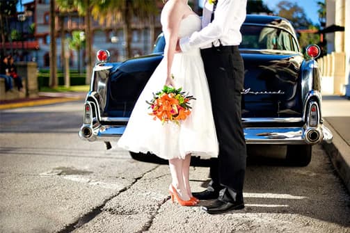 A bride and groom stand closely together, smiling, in front of a vintage car on a sunny street. A bride and groom stand closely together, smiling, in front of a vintage car on a sunny street.