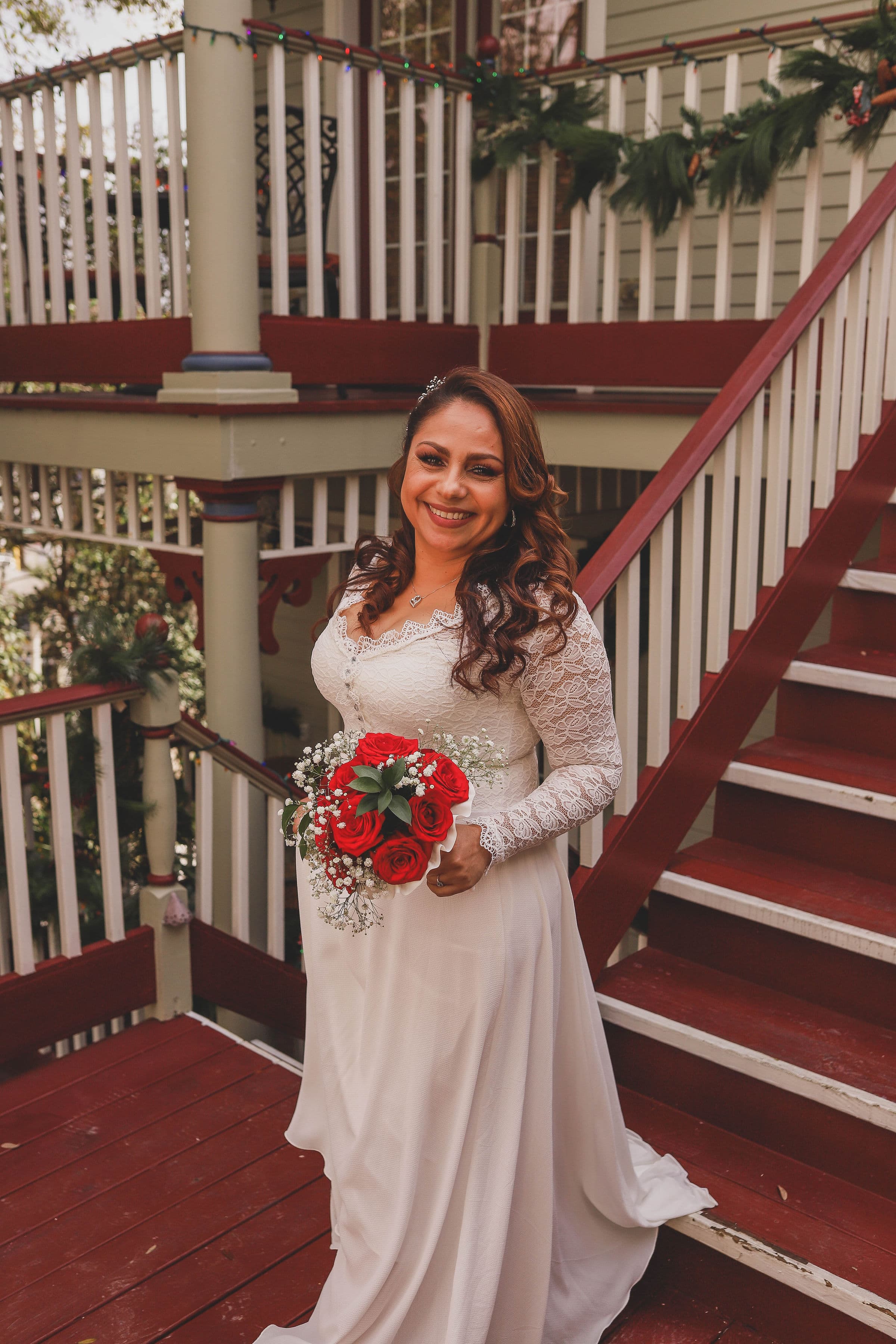 A smiling woman in a white wedding dress holds a bouquet of red roses while standing on a staircase.