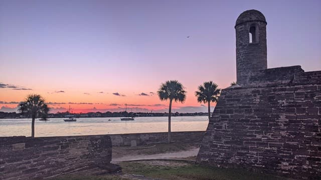 A stone wall with a watchtower overlooks a calm waterway at sunset, framed by palm trees.