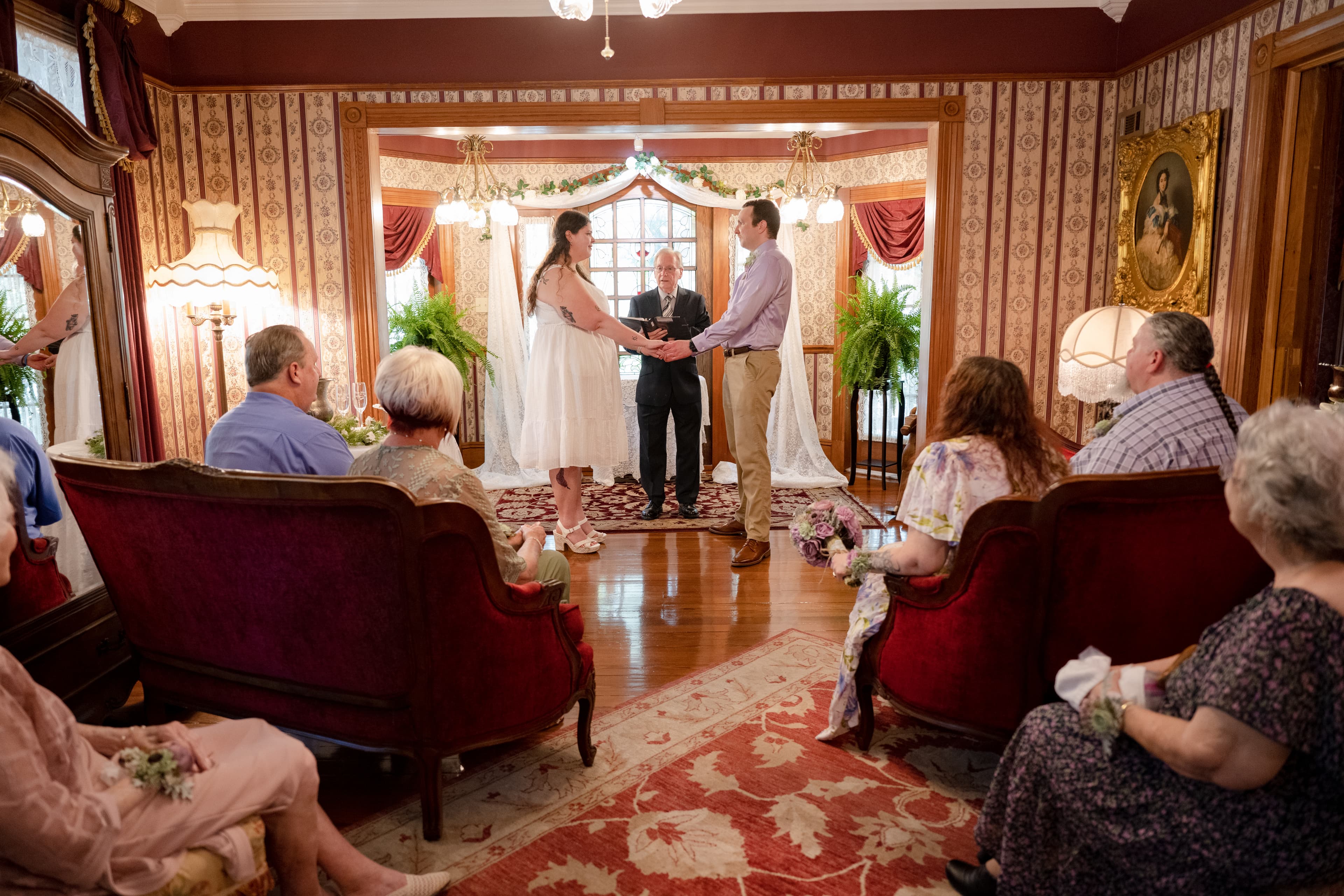A couple stands at the altar exchanging vows in an elegant, intimate living room while guests observe.
